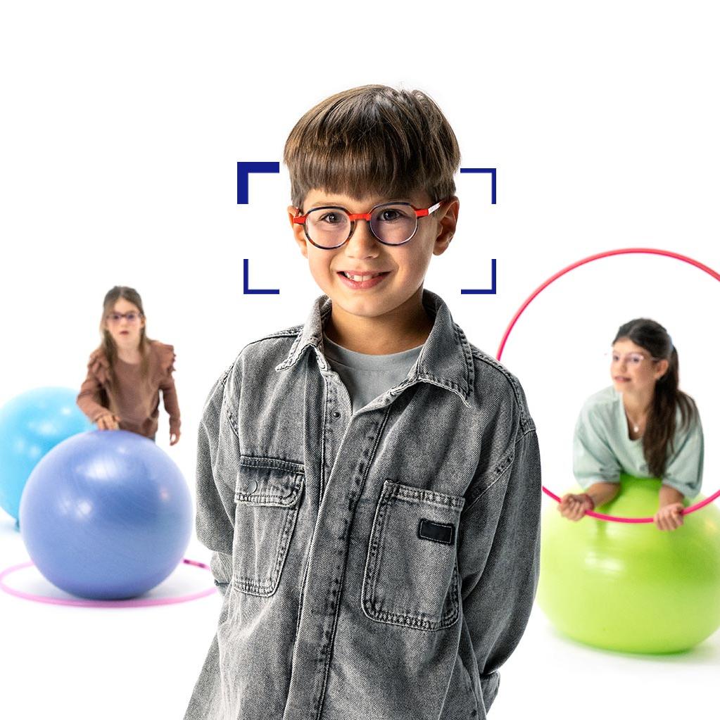 Em primeiro plano vemos um menino de cabelo castanho a usar óculos redondos com lentes ZEISS MyoCare e a sorrir para a câmara. Em segundo plano encontram-se duas meninas a usar lentes ZEISS MyoCare e a brincar com arcos e bolas de ginástica.
