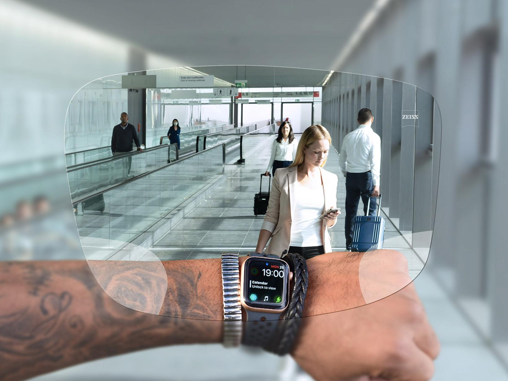 Pessoa a olhar para o relógio através das lentes ZEISS SmartLife Digital. Ao fundo, vê-se uma situação típica de aeroporto, com pessoas a transportarem malas e a caminharem nas escadas rolantes. 