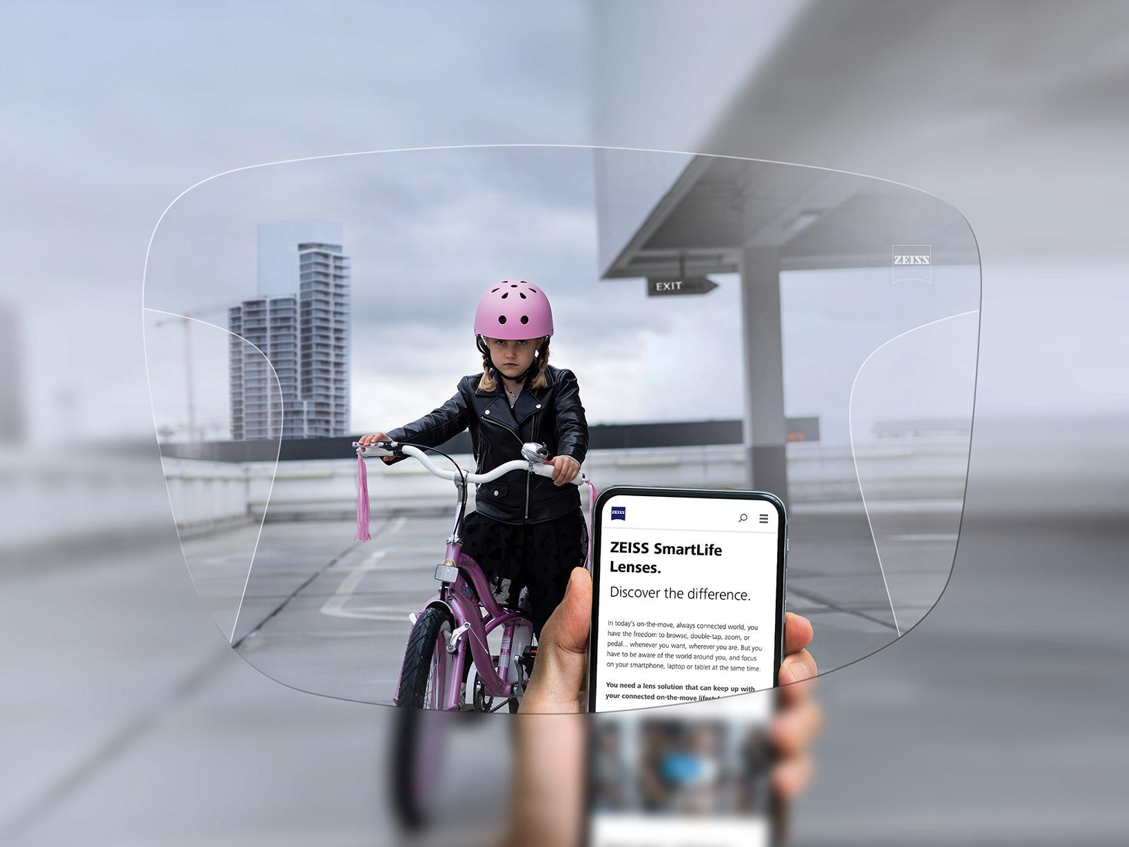 Uma pessoa a olhar para o telemóvel através das lentes ZEISS SmartLife Progressivas Ao fundo, vê-se uma menina de capacete cor-de-rosa a andar de bicicleta. 