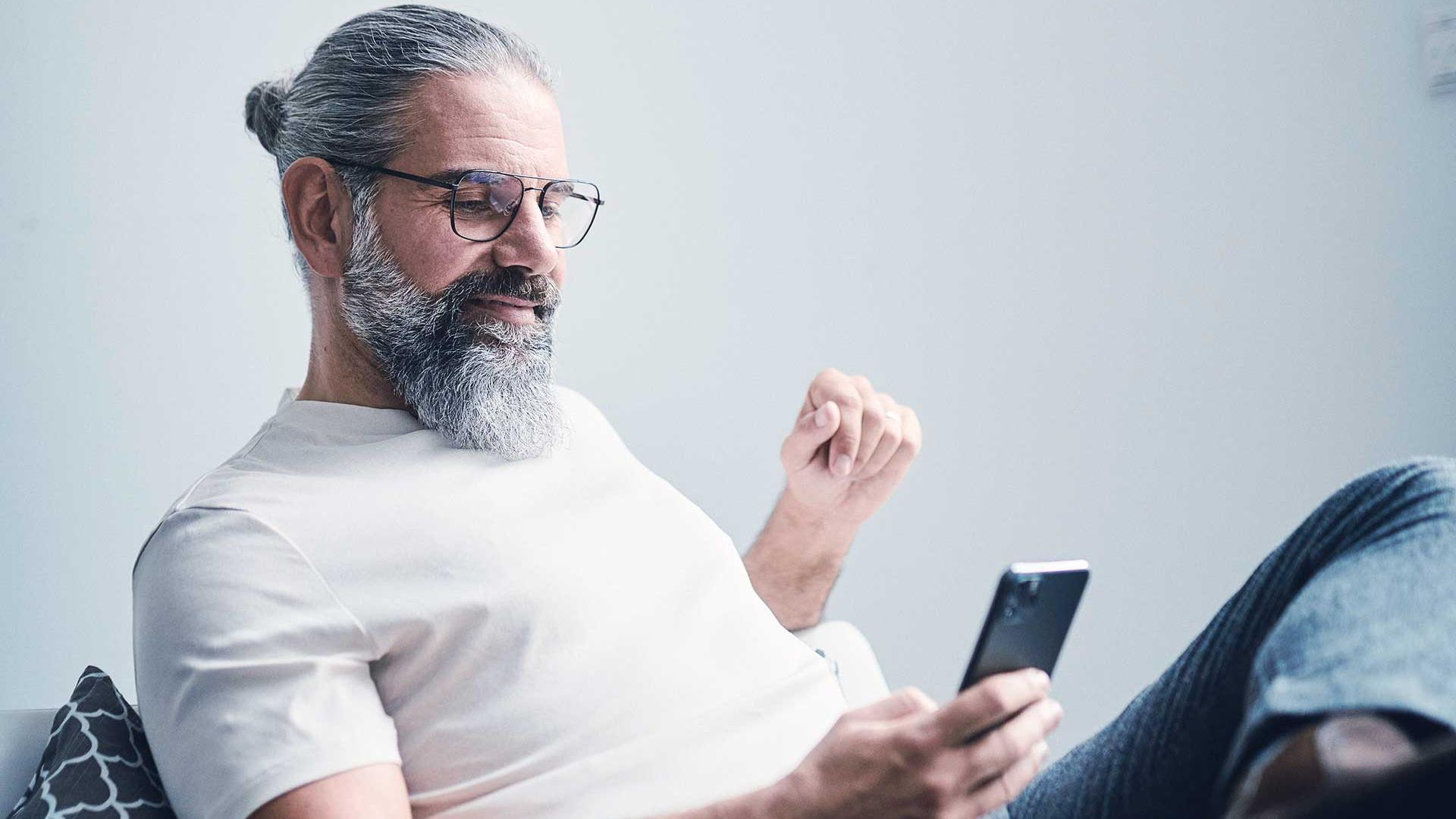 Homem de cabelo grisalho, barba farta e óculos, sentado no sofá a olhar para um smartphone