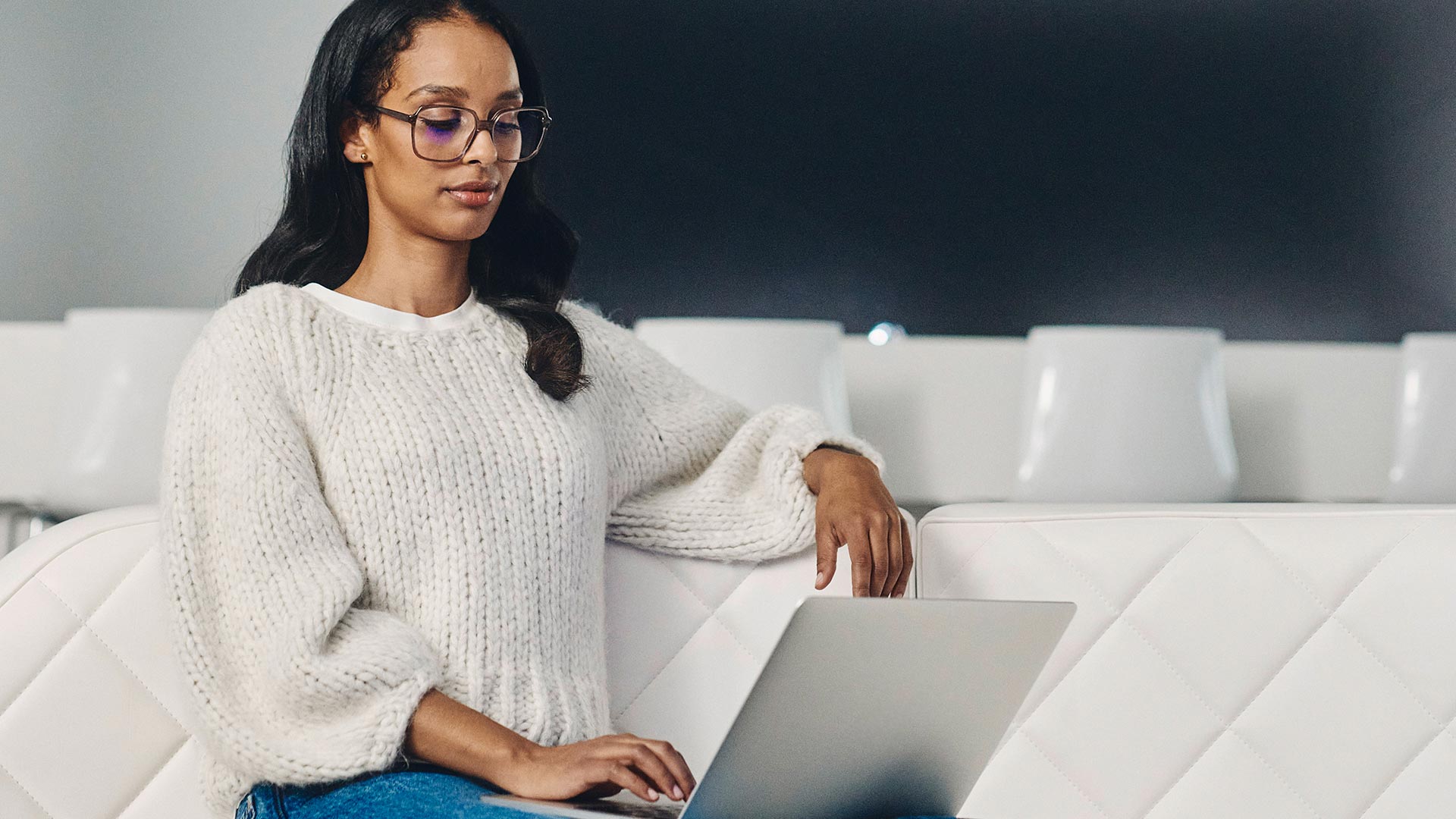 Mulher de cabelo escuro, óculos escuros e camisola branca a olhar para um computador portátil.
