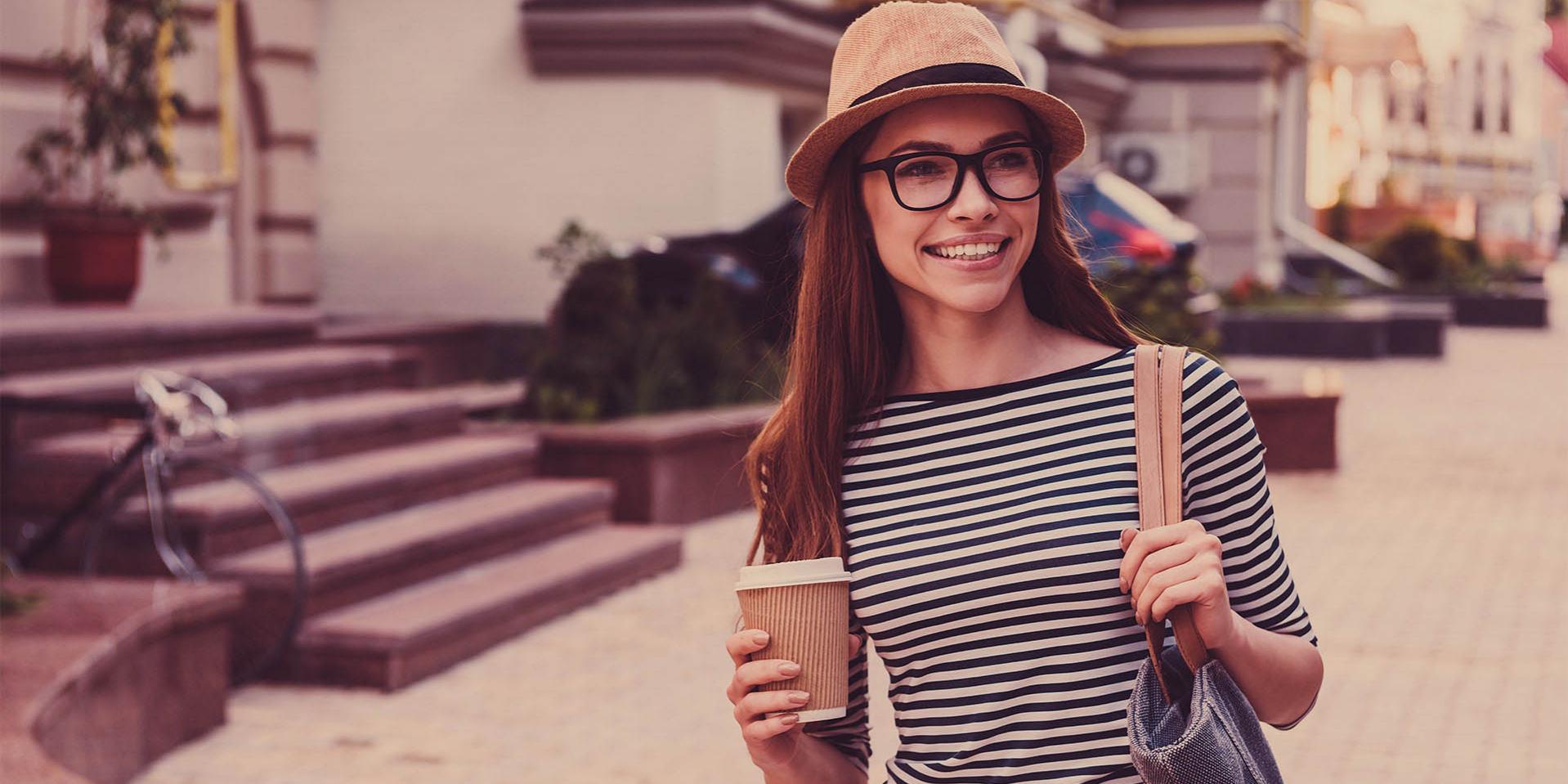 Uma jovem com um chapéu e óculos tem uma caneca de café (lado direito) e uma bolsa no ombro esquerdo