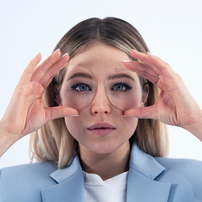 Jovem loira a segurar umas lentes em frente aos olhos para mostrar a sua boa aparência, sem efeitos que distorcem os olhos, graças às lentes monofocais ZEISS ClearView.