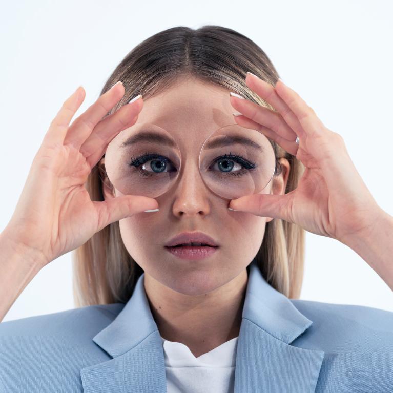 Jovem loira a segurar umas lentes em frente aos olhos para mostrar o efeito de fundo de garrafa provocado pelas lentes grossas.