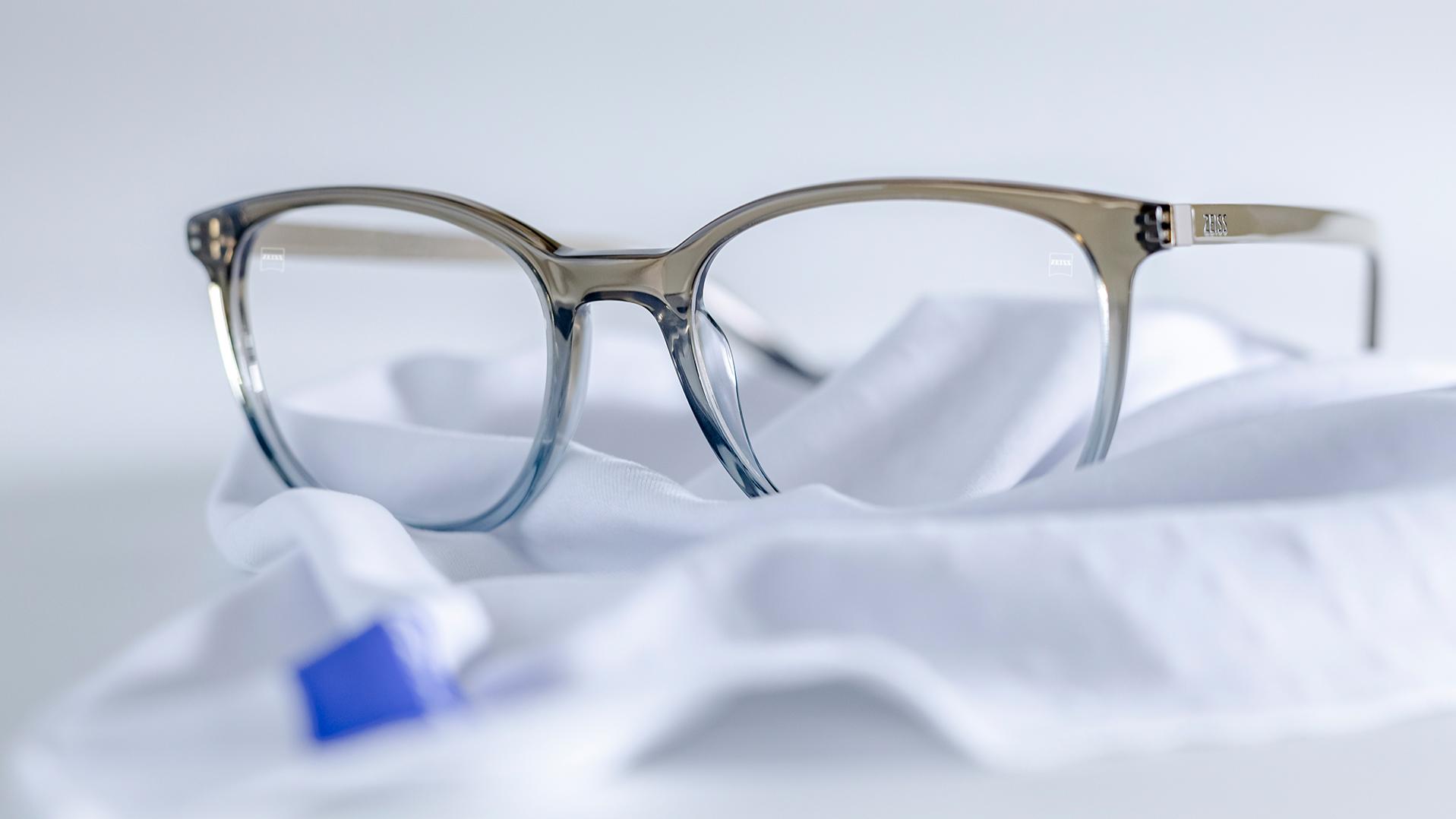 Um par de óculos com armação azul-acinzentada e lentes ZEISS com revestimento DuraVision® sobre um pano de microfibra branco.