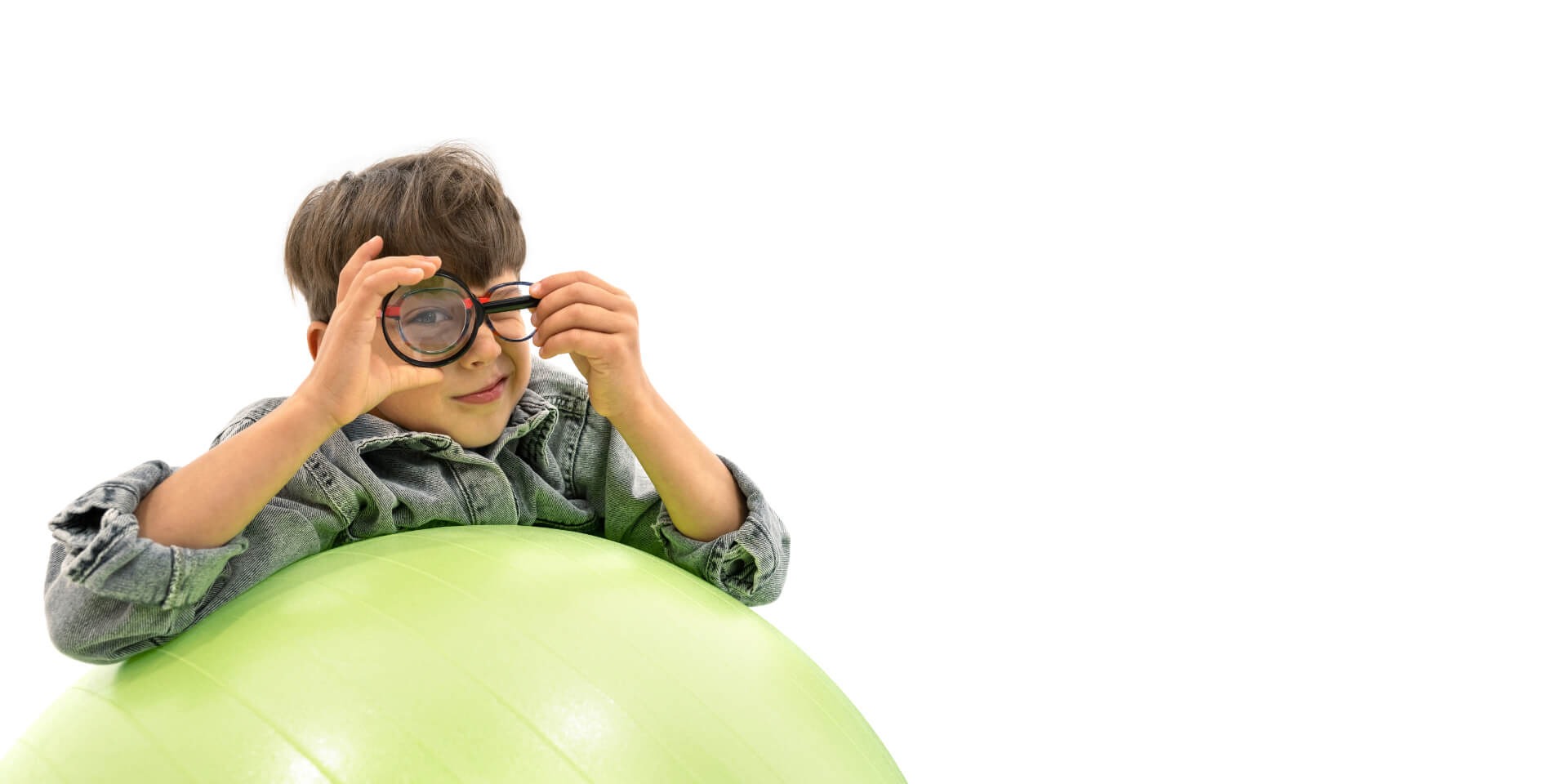 Menino com lentes ZEISS para a gestão da miopia apoiado numa bola de ginástica e a segurar uma lupa à frente do olho.