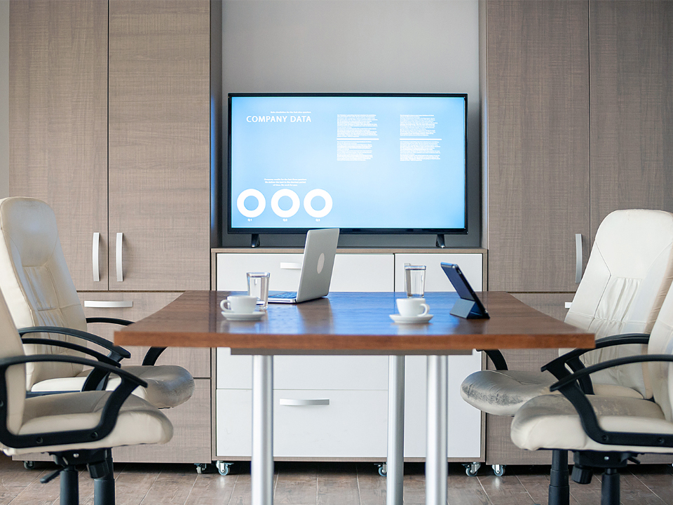 Vista da secretária de um gabinete executivo. Um monitor está montado na parede a alguns metros de distância. Há uma pequena mesa de reuniões com cadeiras de escritório e computadores portáteis em cima da mesa.