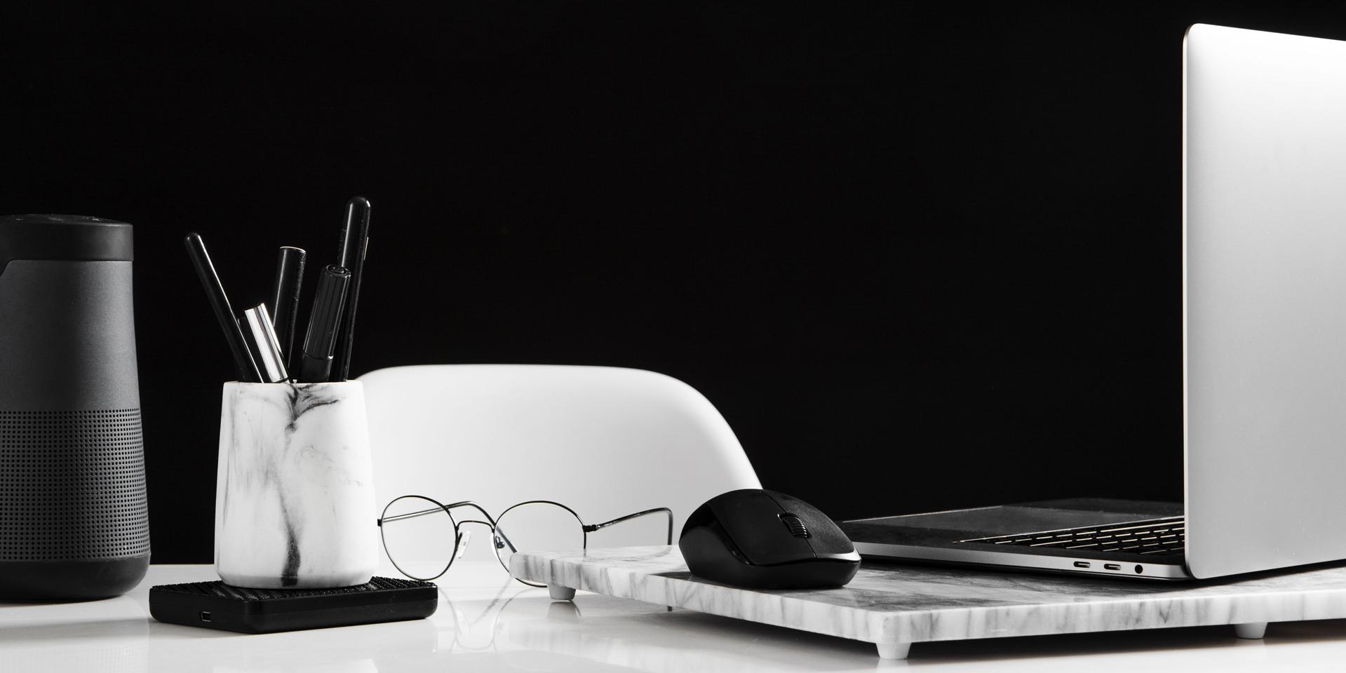 Espaço de trabalho arrumado com um computador portátil aberto do lado direito e um rato. Em frente, está um par de óculos. No lado esquerdo da imagem, encontra-se um copo com canetas.