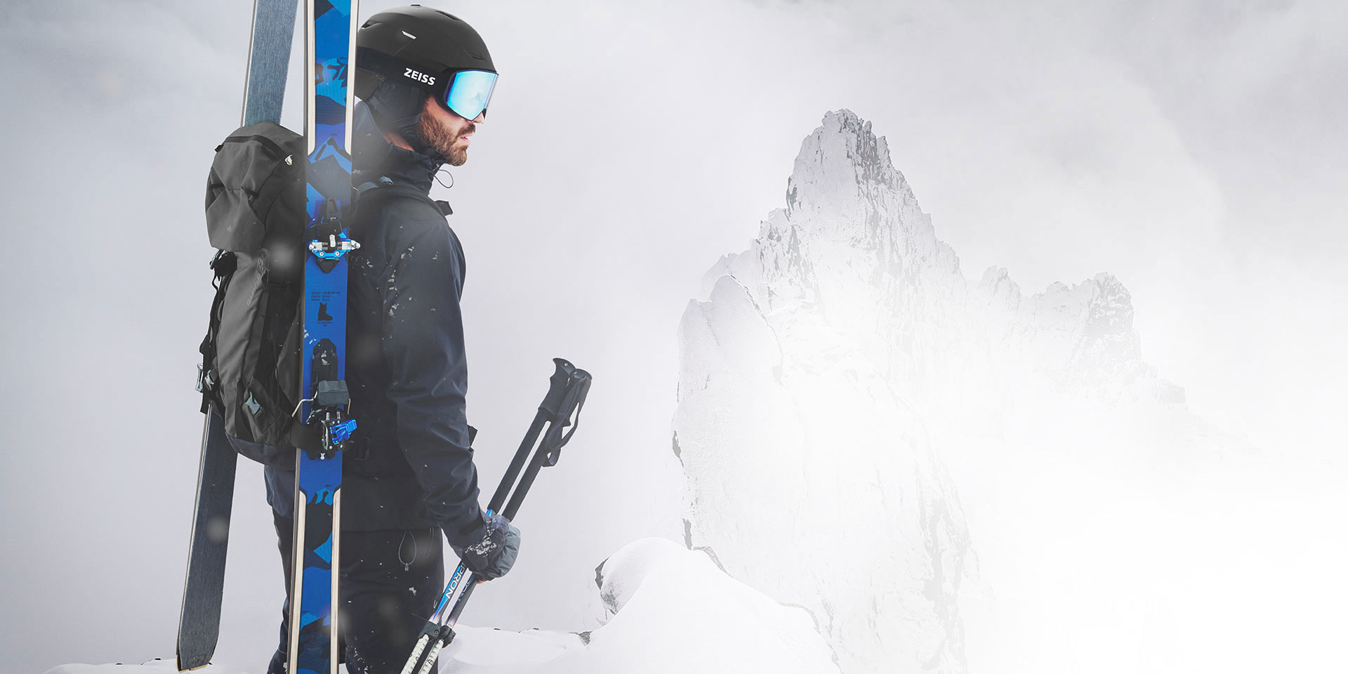 Homem a usar capacete preto e máscara ZEISS para a neve com lente azul espelhada, a transportar os esquis e uma mochila às costas e a segurar os bastões de esqui com a mão direita. Está de pé no cimo de uma montanha.