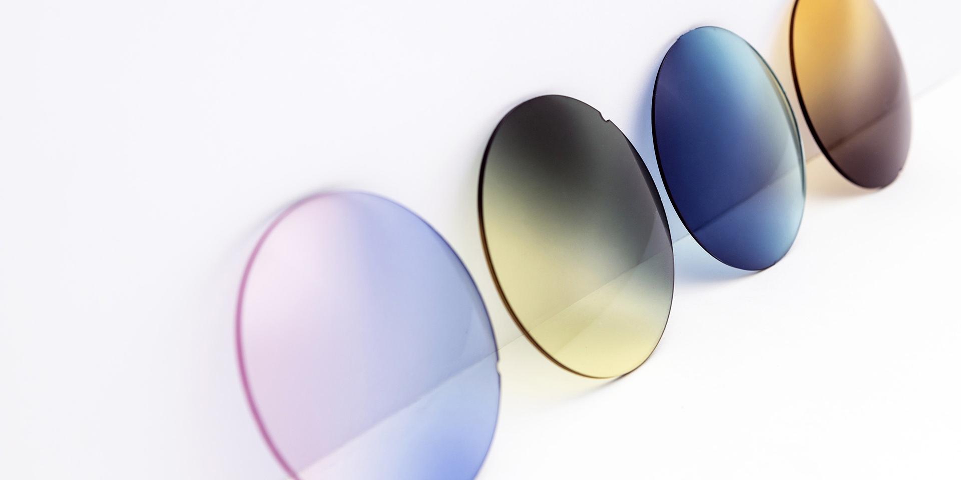 Lentes para óculos de sol de cores diferentes encostadas a uma superfície branca: degradês rosa-púrpura, amarelo-cinzento, azul e castanho.