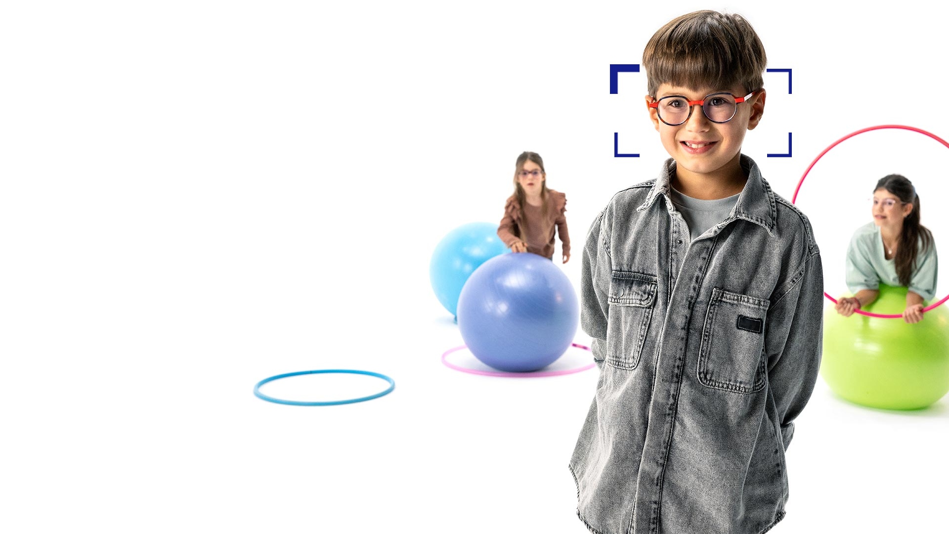 Em primeiro plano vemos um menino de cabelo castanho a usar óculos redondos com lentes ZEISS MyoCare e a sorrir para a câmara. Em segundo plano encontram-se duas meninas a usar lentes ZEISS MyoCare e a brincar com arcos e bolas de ginástica.
